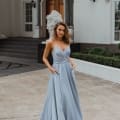 A woman stands outdoors in front of a grand building reminiscent of the elegant architecture found on the Sunshine Coast, wearing the MONROE PO891, a long, light blue, sleeveless gown with thin straps and pockets. She has one hand in a pocket and the other hanging by her side. The building features pillars, windows, and ornamental design. Angels Formal Wear