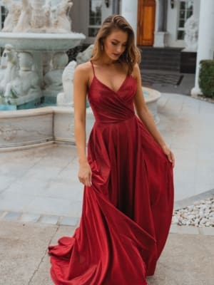 A woman in a flowing MONROE PO891 gown stands on a paved pathway in front of an ornate fountain. The dress features thin straps and a fitted bodice with a full skirt. The scene captures the elegance reminiscent of the Sunshine Coast, with a backdrop featuring a white building adorned with tall columns and an arched wooden door. Angels Formal Wear