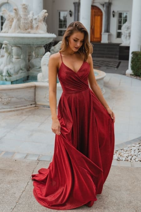 A woman in a flowing MONROE PO891 gown stands on a paved pathway in front of an ornate fountain. The dress features thin straps and a fitted bodice with a full skirt. The scene captures the elegance reminiscent of the Sunshine Coast, with a backdrop featuring a white building adorned with tall columns and an arched wooden door. Angels Formal Wear