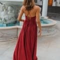 A woman with wavy brown hair stands with her back to the camera, wearing the MONROE PO891, a flowing red dress with a crisscrossed back. She is near a decorative fountain adorned with sculpted figures, situated in front of a building featuring pillars and a wooden door, reminiscent of the elegant architecture on the Sunshine Coast. Angels Formal Wear