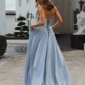 A woman in a flowing MONROE PO891 light blue backless gown stands outdoors in front of a grand white building. The dress features a lace-up back design, and the woman is looking away from the camera while holding the sides of her dress. Set against lush greenery, it feels like a scene straight from the Sunshine Coast. Angels Formal Wear