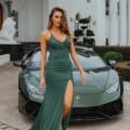 A woman in a green evening gown with a high slit stands confidently in front of a green luxury sports car parked by an elegant white building on the Sunshine Coast. She has long hair and is wearing high heels. The building boasts tall columns and manicured shrubs, exuding timeless charm. Angels Formal Wear