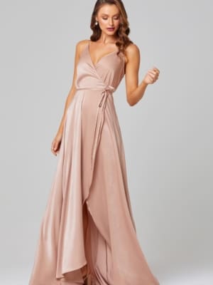 A woman with long, wavy brown hair is wearing the JITHYA TO853, a silky, blush-colored, sleeveless wrap dress that ties at the waist. She stands looking downwards, one hand slightly raised as if adjusting a strap or holding an object. The background is a plain, light gray. Buy wedding dresses online in Australia for similar styles. Angels Formal Wear