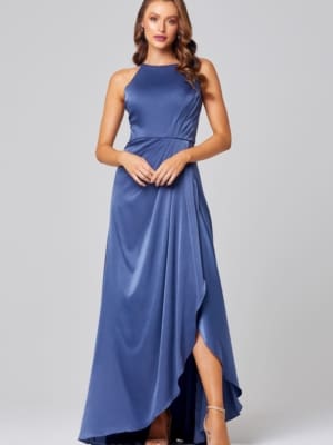 A woman is modeling the CHELSEA TO854, a sleeveless, floor-length blue evening gown featuring a high neckline, pleated detailing on the bodice, and an asymmetrical front slit. Her long, wavy hair complements her high-heeled sandals perfectly. The plain gray background further accentuates this stunning look. Buy Wedding Dresses Online Australia for more options like this. Angels Formal Wear