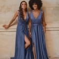 Two women in matching floor-length, slate blue dresses stand against a beige textured wall. The dresses feature plunging necklines and thigh-high slits. The woman on the left has long, wavy hair and has one leg extended. The woman on the right, wearing white heels, showcases her curly hair in this chic Formal Wear Sunshine Coast ensemble. Angels Formal Wear