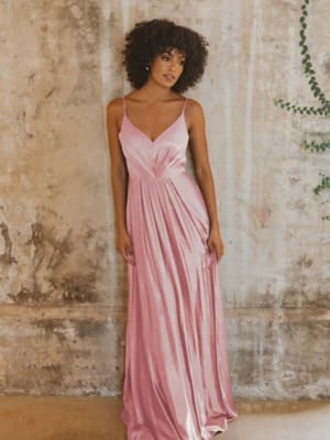 A woman with curly hair is wearing a long, flowing pink dress with spaghetti straps. She is standing against a textured, beige wall with a slightly weathered appearance, and some green plant vines are visible in the upper right corner. For similar formal dresses on the Sunshine Coast, buy online now. Angels Formal Wear