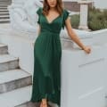 A woman wearing a green dress from the Sunshine Coast Dress Shop stands confidently with one hand resting on a white stone railing and the other on her hip. She is on a staircase next to a classical-style statue. The surroundings are elegant, with a backdrop of a white building and greenery. Angels Formal Wear