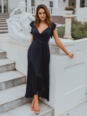 A woman in a BRIGHTON TO865 dress from Sunshine Coast Dress Shop, characterized by its navy blue hue, V-neck design, and ruffled sleeves, stands on a staircase beside a large white statue. She gazes at the camera with a neutral expression. The background features a white balustrade and potted plants. Angels Formal Wear