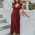 A woman in a burgundy bridesmaid dress poses on a set of outdoor stairs next to a white stone sculpture of a reclining woman. She has light brown hair, wears strappy sandals, and stands in front of a white building with columns, capturing the elegance of the Sunshine Coast setting. Angels Formal Wear