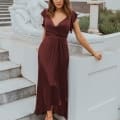 A woman in a burgundy dress, possibly one of the chic bridesmaid dresses from the Sunshine Coast, stands on a stone staircase next to a white marble statue. She leans against a low wall with greenery and a planter in the background, enhancing the elegant outdoor setting with architectural details. Angels Formal Wear