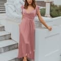 A woman in a pink bridesmaid dress is standing on the steps of an elegant building. She is gently leaning against a white stone sculpture of a seated figure. The building, reminiscent of those on the Sunshine Coast, has white pillars, arched windows, and potted plants. Her hair is styled in loose waves. Angels Formal Wear