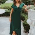 A woman in a green wrap dress stands outdoors next to a decorative urn and well-manicured greenery on the Sunshine Coast. She has shoulder-length hair and wears beige strappy heels. A fountain and garden can be seen in the background, reminiscent of charming bridesmaid dresses settings. Angels Formal Wear