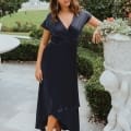 A woman is standing on a stone path in a lush garden on the Sunshine Coast, wearing a dark blue wrap dress and beige high-heeled sandals. Her hand rests on a decorative stone planter featuring cherubic figures. There's a white table and a fountain visible in the background. Angels Formal Wear