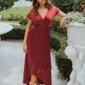 A woman stands outdoors in a garden on the Sunshine Coast, dressed in a burgundy, short-sleeved, asymmetrical dress with a V-neckline. She poses near a white decorative stone planter with sculpted cherubs, the background featuring manicured bushes and a stone fountain—a perfect setting for bridesmaid dresses. Angels Formal Wear