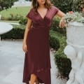 A woman in a burgundy bridesmaid dress stands outdoors near a garden statue, surrounded by greenery. She poses with one hand on the statue and the other hanging by her side. The setting includes neatly trimmed bushes and a white fountain in the background, reminiscent of a scenic spot on the Sunshine Coast. Angels Formal Wear