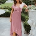 A woman with shoulder-length hair stands outdoors in a garden setting with manicured hedges and a fountain in the background. Wearing a pink wrap dress and beige heels, she rests one hand on a large decorative planter with cherub designs, embodying the elegance of Wedding Dresses Australia. Angels Formal Wear