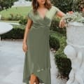 A woman stands outdoors on a stone patio wearing a sage green wrap dress and beige heels. She poses with one hand resting on a large, ornate planter. The background includes neatly trimmed hedges, a white table, and a distant fountain, reminiscent of the elegant settings for Wedding Dresses Australia. Angels Formal Wear