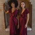 Two women stand side by side in front of an archway set into a textured wall. They both wear the NAPIER TO868, deep burgundy satin bridesmaid dresses featuring short sleeves and asymmetrical hemlines. One woman has curly hair and the other has wavy hair. Both look directly at the camera, capturing the essence of Sunshine Coast elegance. Angels Formal Wear