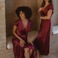 Two women in elegant NAPIER TO868 deep red satin bridesmaid dresses are posing indoors against a textured beige wall. One is seated on a cushioned bench, and the other stands beside her. Both have stylish, wavy hair and appear contemplative. Angels Formal Wear