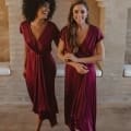 Two women wearing matching NAPIER TO868 burgundy bridesmaid dresses stand barefoot in a room with rustic brick walls. One has curly hair and is smiling widely as she walks, while the other has straight, long hair and smiles softly while holding her right arm with her left hand, reminiscent of a serene Sunshine Coast vibe. Angels Formal Wear