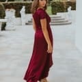 A woman with shoulder-length hair stands outdoors on the Sunshine Coast, wearing a flowing, deep red NAPIER TO868 bridesmaid dress and nude heels. She is turned slightly, looking over her shoulder, with greenery and statues in the background. Angels Formal Wear