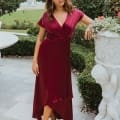 A woman stands outdoors in a garden on the Sunshine Coast, wearing the NAPIER TO868, a flowing maroon bridesmaid dress with short sleeves and a high-low hem. She is leaning slightly on a decorative stone planter with intricate carvings, surrounded by manicured greenery and a fountain in the background. Angels Formal Wear