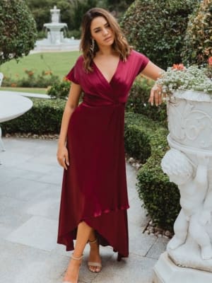 A woman stands outdoors in a garden on the Sunshine Coast, wearing the NAPIER TO868, a flowing maroon bridesmaid dress with short sleeves and a high-low hem. She is leaning slightly on a decorative stone planter with intricate carvings, surrounded by manicured greenery and a fountain in the background. Angels Formal Wear