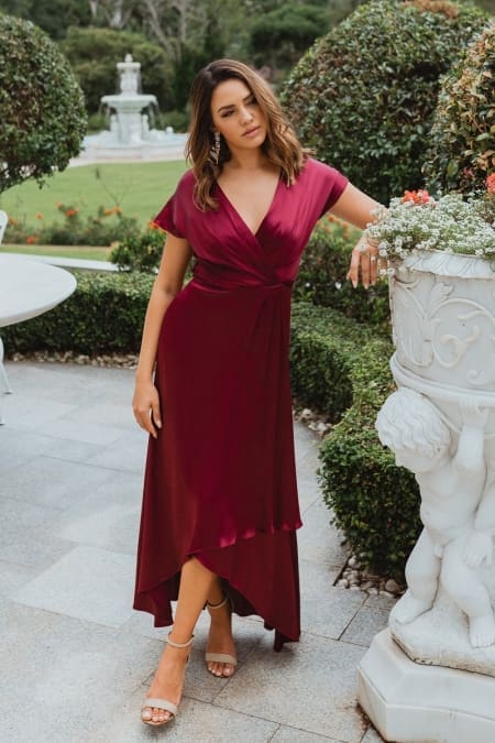 A woman stands outdoors in a garden on the Sunshine Coast, wearing the NAPIER TO868, a flowing maroon bridesmaid dress with short sleeves and a high-low hem. She is leaning slightly on a decorative stone planter with intricate carvings, surrounded by manicured greenery and a fountain in the background. Angels Formal Wear