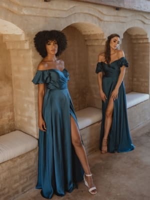 Two women wearing elegant LAGOS TO873 off-the-shoulder gowns in teal pose in a rustic setting. The floor-length dresses, available at the Sunshine Coast Dress Shop, feature thigh-high slits. One woman stands against a wall with a natural afro hairstyle, while the other leans slightly forward toward the camera. Angels Formal Wear