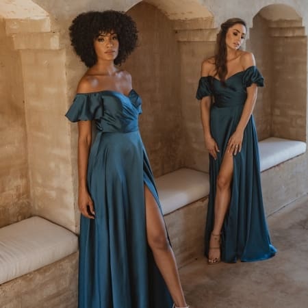 Two women wearing elegant LAGOS TO873 off-the-shoulder gowns in teal pose in a rustic setting. The floor-length dresses, available at the Sunshine Coast Dress Shop, feature thigh-high slits. One woman stands against a wall with a natural afro hairstyle, while the other leans slightly forward toward the camera. Angels Formal Wear