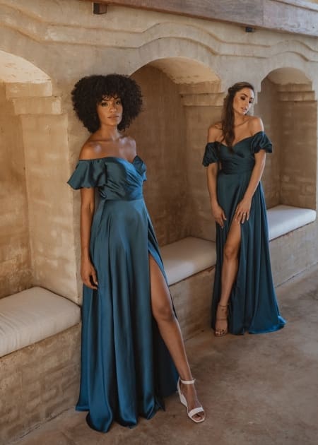 Two women wearing elegant LAGOS TO873 off-the-shoulder gowns in teal pose in a rustic setting. The floor-length dresses, available at the Sunshine Coast Dress Shop, feature thigh-high slits. One woman stands against a wall with a natural afro hairstyle, while the other leans slightly forward toward the camera. Angels Formal Wear