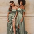 Two women in green off-the-shoulder satin formal dresses stand side by side, smiling. Both dresses feature thigh-high slits. One woman has long, wavy hair, and the other has curly hair. The background is a neutral, textured wall. Discover these exquisite styles and buy online from Formal Dresses Sunshine Coast. Angels Formal Wear