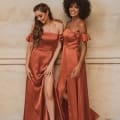 Two women pose together in matching off-the-shoulder, copper-colored satin formal dresses with thigh-high slits. One has curly hair, and the other has wavy hair pulled to the side. They stand against a textured beige wall, smiling and looking at each other. Buy online for elegant looks from Sunshine Coast. Angels Formal Wear