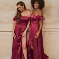 Two women stand side by side in matching OSAKA TO874 off-the-shoulder burgundy gowns with thigh-high slits. One has long, wavy hair; the other has curly hair. Smiling and looking down, they pose against a beige textured wall. Discover similar formal dresses from the Sunshine Coast and buy online today! Angels Formal Wear