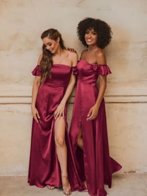 Two women stand side by side in matching OSAKA TO874 off-the-shoulder burgundy gowns with thigh-high slits. One has long, wavy hair; the other has curly hair. Smiling and looking down, they pose against a beige textured wall. Discover similar formal dresses from the Sunshine Coast and buy online today! Angels Formal Wear