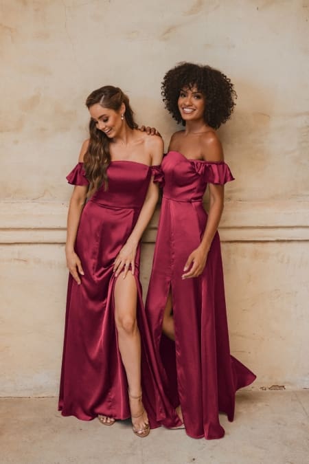 Two women stand side by side in matching OSAKA TO874 off-the-shoulder burgundy gowns with thigh-high slits. One has long, wavy hair; the other has curly hair. Smiling and looking down, they pose against a beige textured wall. Discover similar formal dresses from the Sunshine Coast and buy online today! Angels Formal Wear