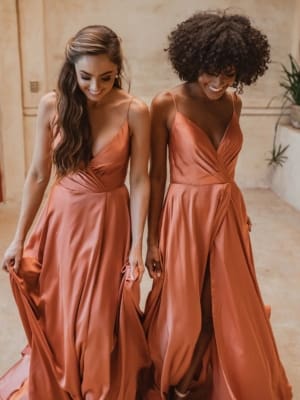 Two women in flowing, floor-length SUVA TO875 dresses smile while looking down. The formal wear features sleeveless designs with deep V-necks and gathered waists, creating a gentle drape. They stand on a light-colored floor with a soft, neutral-toned background, reminiscent of Sunshine Coast elegance. Angels Formal Wear