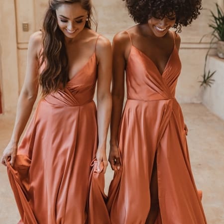 Two women in flowing, floor-length SUVA TO875 dresses smile while looking down. The formal wear features sleeveless designs with deep V-necks and gathered waists, creating a gentle drape. They stand on a light-colored floor with a soft, neutral-toned background, reminiscent of Sunshine Coast elegance. Angels Formal Wear