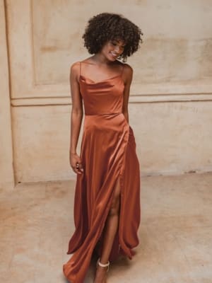 A woman with curly hair is wearing a long, elegant, reddish-orange satin dress with thin straps and a thigh-high slit. She is standing in a beige, minimalist room and is smiling while looking slightly downwards. Her white sandals complete the perfect formal wear look for an event on the Sunshine Coast. Angels Formal Wear