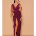 A woman stands against a beige background wearing the DEMI NBM1001, a sleeveless, deep V-neck burgundy gown with a high thigh slit, revealing one leg. She has long, wavy dark hair and wears strappy heels, looking downwards with one hand resting on her hip. Angels Formal Wear