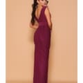 A woman stands with her back turned, showcasing a DEMI NBM1001 floor-length, sleeveless, deep maroon gown with a V-shaped back and draped detailing. Her long, dark hair is styled in loose waves as she poses against a simple beige background. Angels Formal Wear