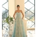 A woman stands in an elegant room wearing a light blue strapless lace gown with a sweetheart neckline, available at Formal Wear Shop Sunshine Coast. The EMBER NC1057 complements her style perfectly. She has her hair styled in a high bun and rests her hands on a white column behind her. Large windows with a grid pattern are in the background, letting in natural light. Angels Formal Wear