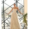 A woman wearing a strapless white lace gown poses on an indoor balcony with ornate black iron railings and large windows. The sunlight streams through the windows, illuminating her dress. This stunning scene could easily be from an EMBER NC1057 on the Sunshine Coast. Angels Formal Wear