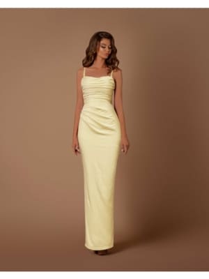 A woman stands in front of a plain brown background, wearing a form-fitting, floor-length, yellow satin dress with spaghetti straps and EMMA NBM1024 ruching detail on the bodice. Her long, wavy hair cascades over her shoulders. Angels Formal Wear