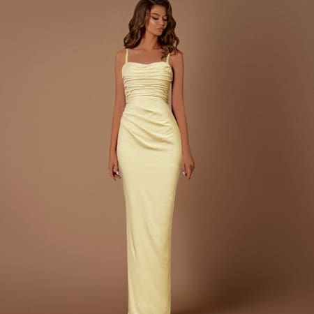 A woman stands in front of a plain brown background, wearing a form-fitting, floor-length, yellow satin dress with spaghetti straps and EMMA NBM1024 ruching detail on the bodice. Her long, wavy hair cascades over her shoulders. Angels Formal Wear