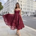 A woman stands on a city sidewalk wearing a sleeveless, burgundy lace dress with a flared, mid-length skirt, the PIXIE JX5031. She is holding the sides of her dress out slightly. The street features white buildings with multiple windows, and there are a few parked cars in the background. Angels Formal Wear