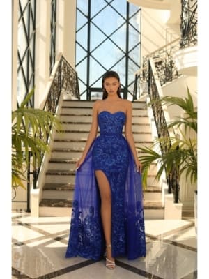 A woman stands on a grand marble staircase lined with potted plants, wearing the STELLA NC1003 with a high slit and flowing train. She has long, straight hair and wears silver high-heeled sandals. The setting features large windows letting in natural light. Angels Formal Wear