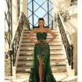 A woman stands confidently at the bottom of an elegant staircase adorned with intricate wrought-iron railings. She is wearing a STELLA NC1003, paired with high heels. The background features large windows with a geometric design. Angels Formal Wear