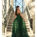 A woman wearing a long green STELLA NC1003 formal gown stands on an elegant staircase with ornate railings. She faces away, holding the sides of her dress, which has intricate detailing at the hem. Tall windows in the background allow natural light to illuminate the scene. Angels Formal Wear