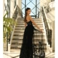 A woman wearing an elegant black strapless STELLA NC1003 gown stands on a grand staircase with intricate iron railings. The gown features a sheer overlay with detailed patterns, and she looks over her shoulder back at the camera. The background showcases large windows. Angels Formal Wear